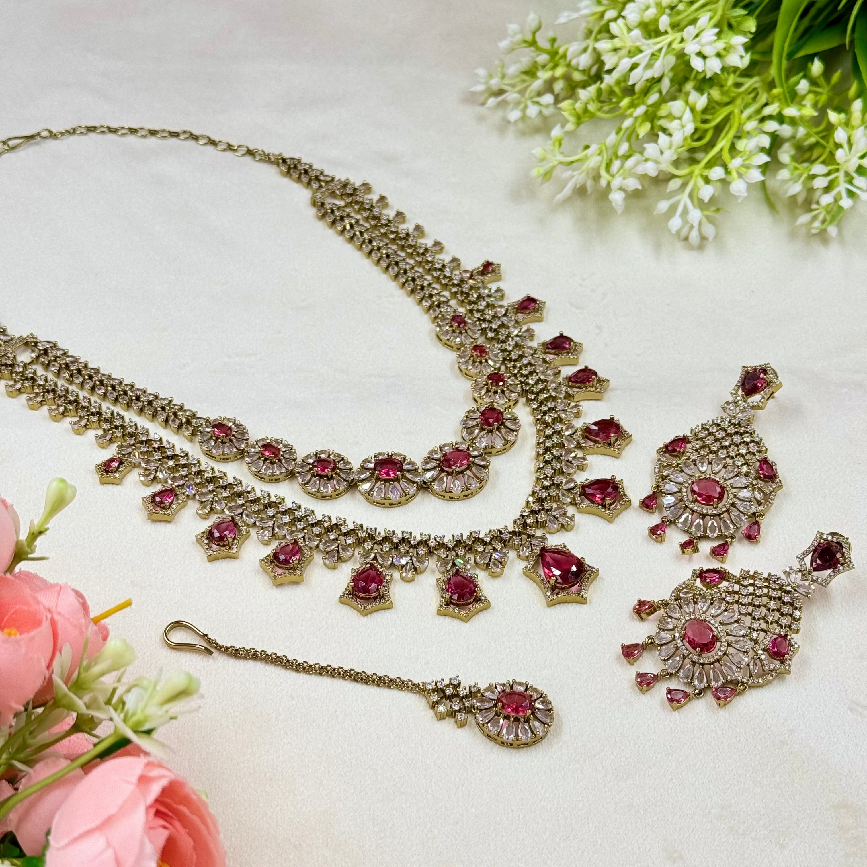 Ornate jewelry set with red gemstones on a light background with flowers.