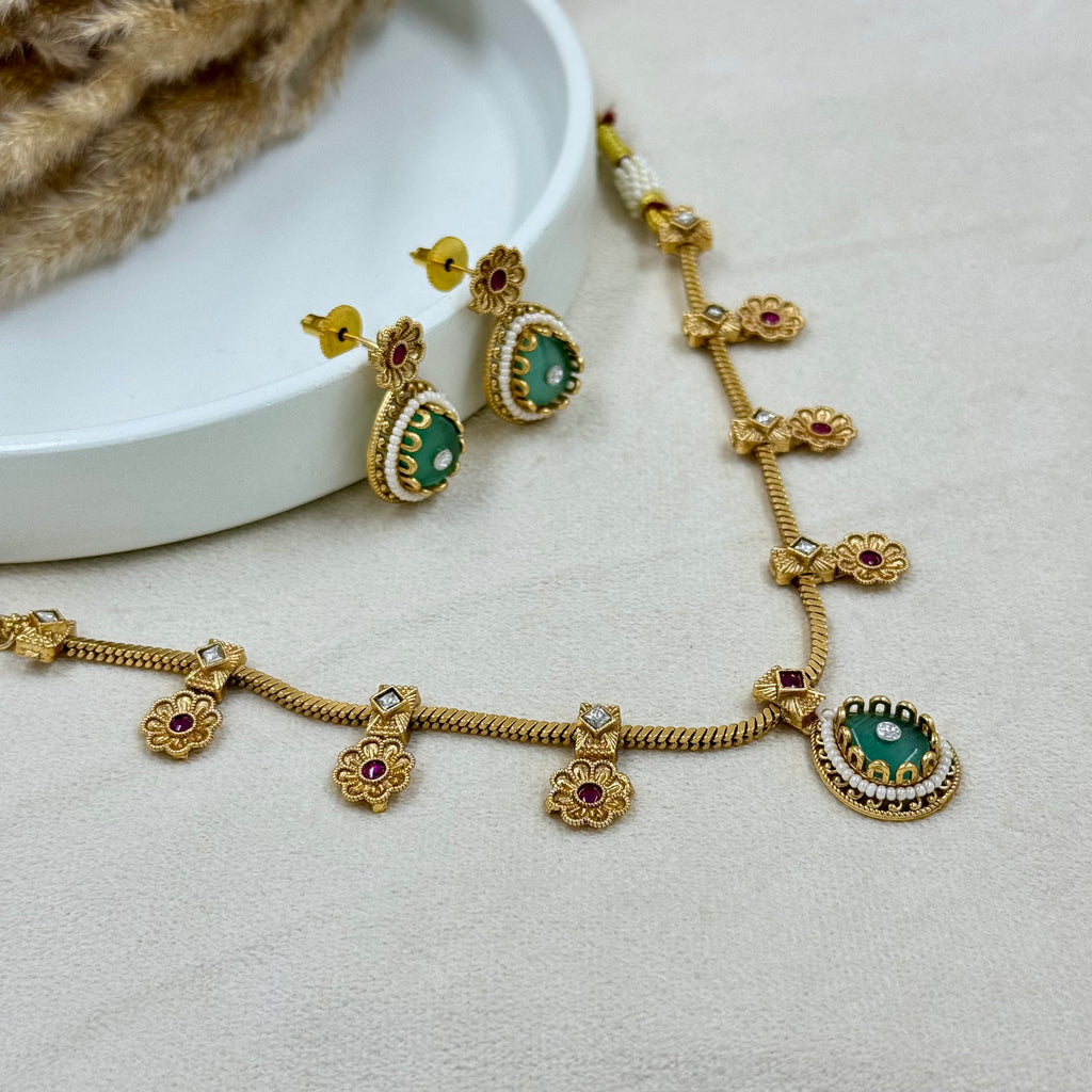 Gold necklace with green gemstones and red stones on a light gray surface.