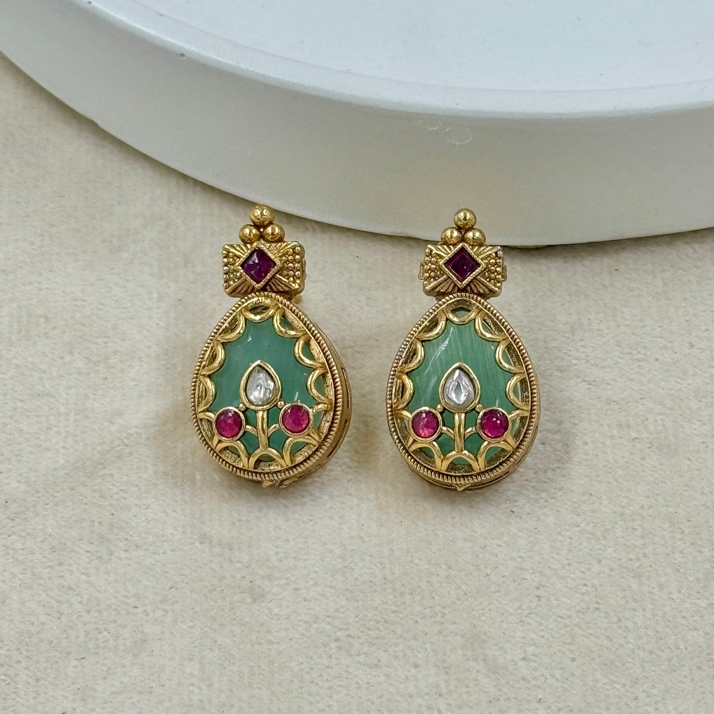 Pair of gold earrings with green and red gemstones on a beige surface
