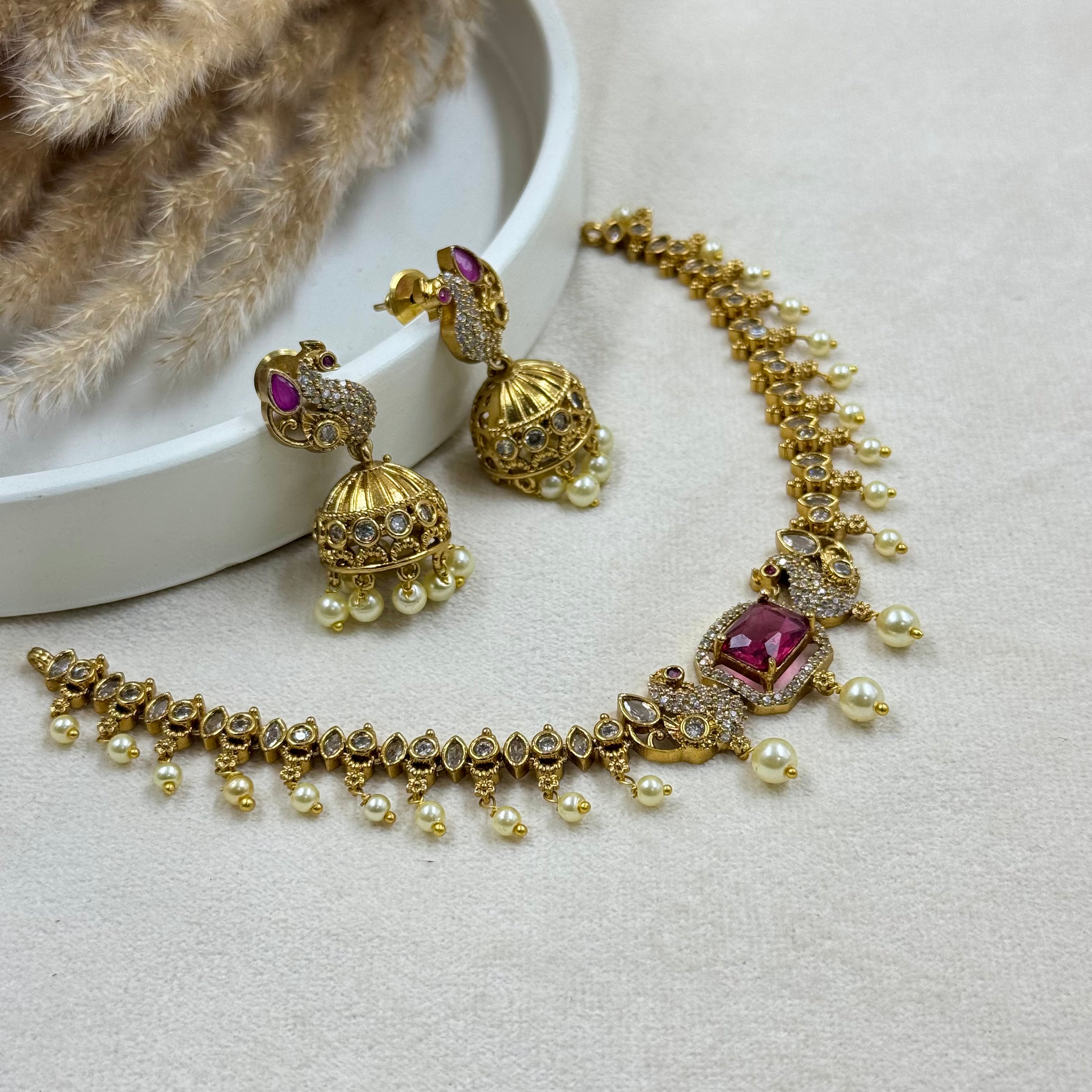 Gold necklace with red gemstones and pearl-like beads, accompanied by matching earrings on a light gray surface.