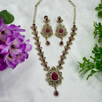Ornate necklace and earrings with red gemstones on a white background with purple flowers and green leaves.