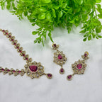 Ornate necklace and earrings with red gemstones on a white background with purple flowers and green leaves.
