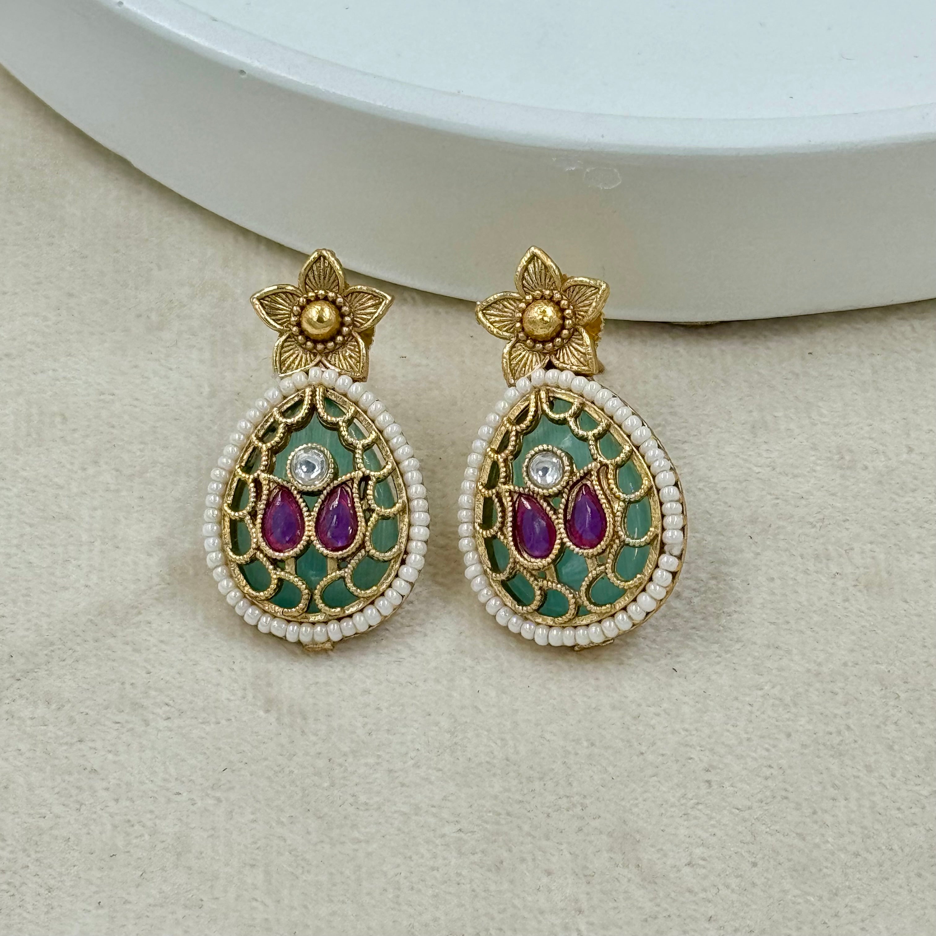 Pair of decorative earrings with gold, purple, and green details on a beige surface.