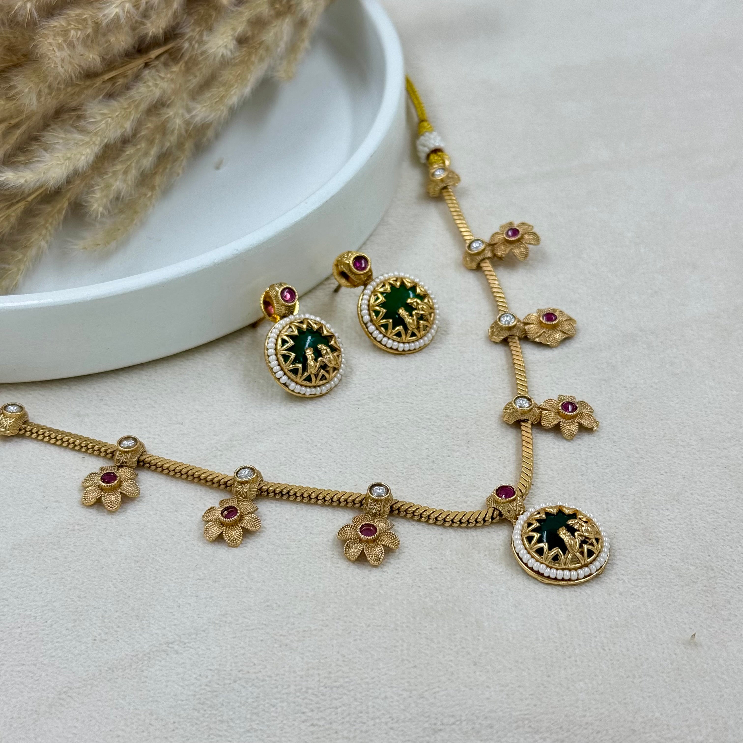 Gold necklace and earrings with green and red stones on a light gray surface.